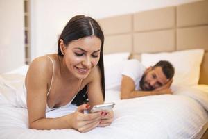 young couple in bed husband frustrated upset and unsatisfied while his internet addict wife is using mobile phone in social network addiction ignoring him in relationship domestic problems photo