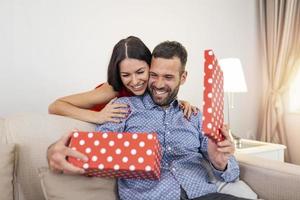 Woman surprising her husband with a gift on the couch at home on Valentine's Day. The beautiful young couple is celebrating at home. A woman is giving her man a gift box photo