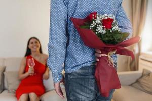 Happy woman looking at her boyfriend holding bouquet of roses behind his back. Handsome man holding flowers behind his back to surprise his girlfriend, romantic happy couples on Valentines day photo