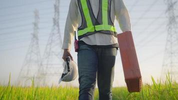 Weißer Helm des Elektroingenieurs und Sicherheitsweste mit Werkzeugkasten, die in der Nähe von Hochspannungsleitungen in Richtung Kraftwerk auf dem Feld gehen. video