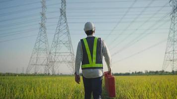 Electrical engineer White helmet and safety carrying toolbox vest walking near high voltage electrical lines towards power station on the field. video