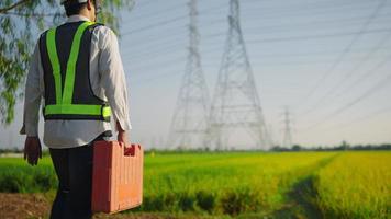 Weißer Helm des Elektroingenieurs und Sicherheitsweste mit Werkzeugkasten, die in der Nähe von Hochspannungsleitungen in Richtung Kraftwerk auf dem Feld gehen. video