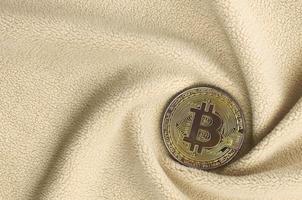 The golden bitcoin lies on a blanket made of soft and fluffy light orange fleece fabric with a large number of relief folds. The shape of the folds resembles a fan from a video card cooler photo