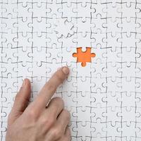 The texture of a white jigsaw puzzle in the assembled state with one missing element, forming an orange space, pointed to by the finger of the male hand photo