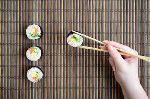 A hand with chopsticks holds a sushi roll on a bamboo straw serwing mat background. Traditional Asian food photo