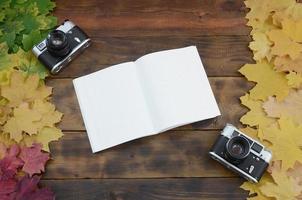 An open notebook and two old cameras among a set of yellowing fallen autumn leaves on a background surface of natural wooden boards of dark brown color photo