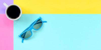 pequeña taza de café con leche y gafas de sol azules sobre fondo de textura de papel de colores azul pastel, amarillo, violeta y rosa de moda en un concepto mínimo foto