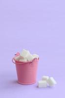 A miniature pink bucket filled with cubes of sugar lies on a purple pastel background. Minimal concept photo