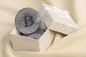 The silver bitcoin lies in a small orange gift box with a small bow on a blanket made of soft and fluffy light orange fleece fabric with a large number of relief folds photo