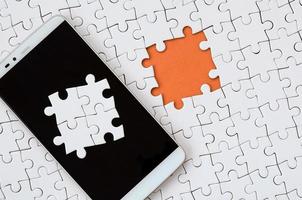 A modern big smartphone with several puzzle elements on the touch screen lies on a white jigsaw puzzle in an assembled state with missing elements photo