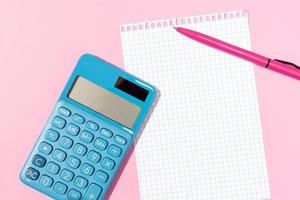 Flat lay of a calculator, blank grid paper notebook and pen on pink background photo