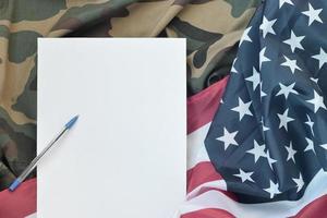 Blank paper lies on United States of America flag and folded military uniform jacket. Military symbols conceptual background banner and copy space photo