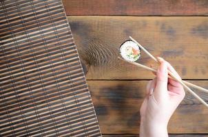 A hand with chopsticks holds a sushi roll on a bamboo straw serwing mat background. Traditional Asian food photo