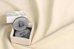 The silver bitcoin lies in a small orange gift box with a small bow on a blanket made of soft and fluffy light orange fleece fabric with a large number of relief folds photo