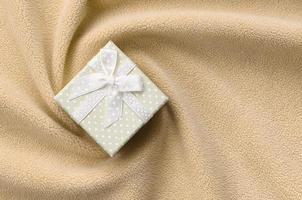 A small gift box in orange with a small bow lies on a blanket of soft and furry light orange fleece fabric with a lot of relief folds. Packing for a gift to your lovely girlfriend photo