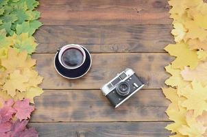 A cup of tea and an old camera among a set of yellowing fallen autumn leaves on a background surface of natural wooden boards of dark brown color photo