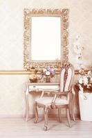 A fragment of a retro interior with a chair and table, on which is a telephone and a vase of flowers. Above the table hangs an empty picture frame for your photo