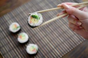 A hand with chopsticks holds a sushi roll on a bamboo straw serwing mat background. Traditional Asian food photo