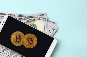 Bitcoins lies on a smartphone and hundred dollar bills on a light blue background photo