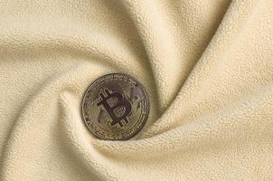 The golden bitcoin lies on a blanket made of soft and fluffy light orange fleece fabric with a large number of relief folds. The shape of the folds resembles a fan from a video card cooler photo