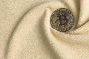 The golden bitcoin lies on a blanket made of soft and fluffy light orange fleece fabric with a large number of relief folds. The shape of the folds resembles a fan from a video card cooler photo