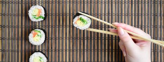 una mano con palillos sostiene un rollo de sushi sobre un fondo de estera de paja de bambú. comida asiática tradicional foto