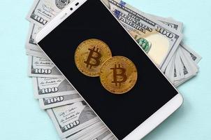 Bitcoins lies on a smartphone and hundred dollar bills on a light blue background photo