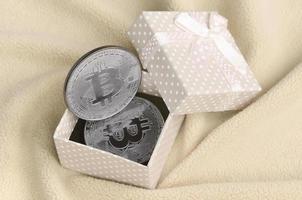 The silver bitcoin lies in a small orange gift box with a small bow on a blanket made of soft and fluffy light orange fleece fabric with a large number of relief folds photo