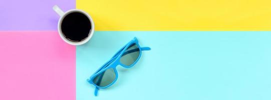 Small white coffee cup and blue sunglasses on texture background of fashion pastel blue, yellow, violet and pink colors paper in minimal concept photo