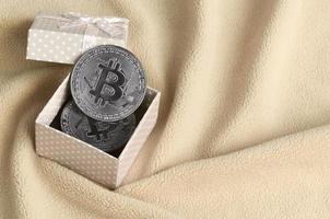 The silver bitcoin lies in a small orange gift box with a small bow on a blanket made of soft and fluffy light orange fleece fabric with a large number of relief folds photo