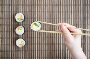 A hand with chopsticks holds a sushi roll on a bamboo straw serwing mat background. Traditional Asian food photo