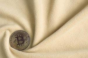 The golden bitcoin lies on a blanket made of soft and fluffy light orange fleece fabric with a large number of relief folds. The shape of the folds resembles a fan from a video card cooler photo