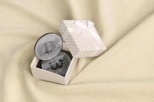 The silver bitcoin lies in a small orange gift box with a small bow on a blanket made of soft and fluffy light orange fleece fabric with a large number of relief folds photo