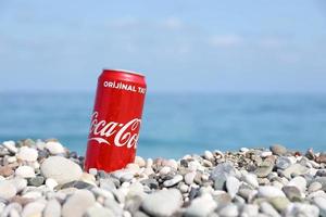 antalya, turquía - 18 de mayo de 2022 la lata roja original de coca cola se encuentra sobre pequeñas piedras redondas cerca de la orilla del mar. coca-cola en playa turca foto