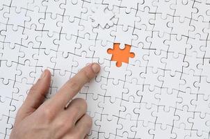 The texture of a white jigsaw puzzle in the assembled state with one missing element, forming an orange space, pointed to by the finger of the male hand photo