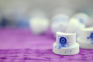 The soiled blue nozzles from the paint sprayer lies on a purple plank on a background of a many dirty spray cans photo