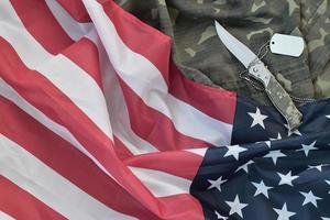 Army dog tag token and knife lies on Old Camouflage uniform and folded United States Flag photo