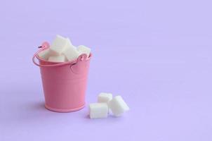 A miniature pink bucket filled with cubes of sugar lies on a purple pastel background. Minimal concept photo