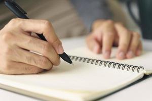 Cerca de joven en manos de tela casual escribiendo en el bloc de notas, cuaderno con bolígrafo sobre la mesa. foto