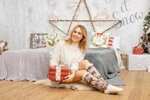 Happy young woman holding a present surrounded by gift boxes sits cross-legged on a camel couch. Christmas gift in the hands of a girl, close-up. photo