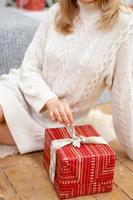 Happy young woman holding a present surrounded by gift boxes sits cross-legged on a camel couch. Christmas gift in the hands of a girl, close-up. photo