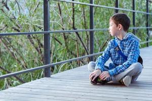 a stylish boy sits on a wooden bridge, among the greenery and looks at the water photo