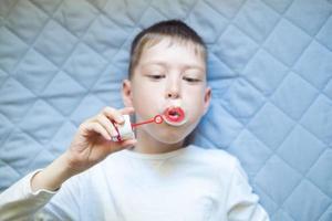 niño acostado en la cama soplando pompas de jabón, primer plano. retrato de un niño con pompas de jabón, vista superior. ejercicios de respiración. foto