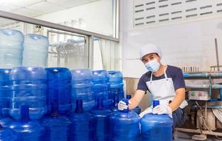 hombre trabajador en ropa de trabajo y con una máscara protectora en la cara trabajando en una fábrica de agua potable revisando galones de agua azul antes del envío. foto