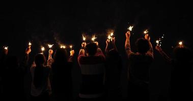 Handaufnahme, Rückansicht, eine Gruppe junger Freunde steht in einer Reihe mit brennenden Wunderkerzen, sie erheben sich und winken am Himmel, um mit funkelnden Händen in der Neujahrspartynacht im Freien zu spielen video
