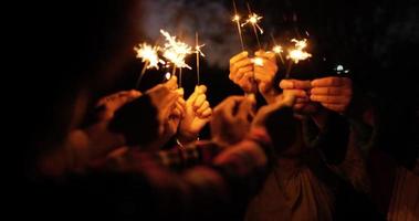 foco seletivo, grupo de jovens amigos segurando estrelinhas queimando fogo, eles estão levantando e acenando para brincar com brilho nas mãos na noite de festa de ano novo ao ar livre video