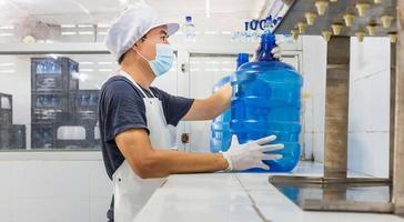 hombre trabajador en ropa de trabajo y con una máscara protectora en la cara trabajando produce agua potable en una fábrica de agua potable limpia. línea de producción de agua potable limpia foto