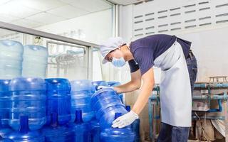 hombre trabajador en ropa de trabajo y con una máscara protectora en la cara trabajando en una fábrica de agua potable revisando galones de agua azul antes del envío. foto