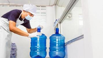 hombre trabajador en ropa de trabajo y con una máscara protectora en la cara trabajando en una fábrica de agua potable revisando galones de agua azul antes del envío. foto