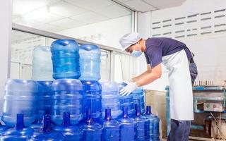 hombre trabajador en ropa de trabajo y con una máscara protectora en la cara trabajando en una fábrica de agua potable revisando galones de agua azul antes del envío. foto